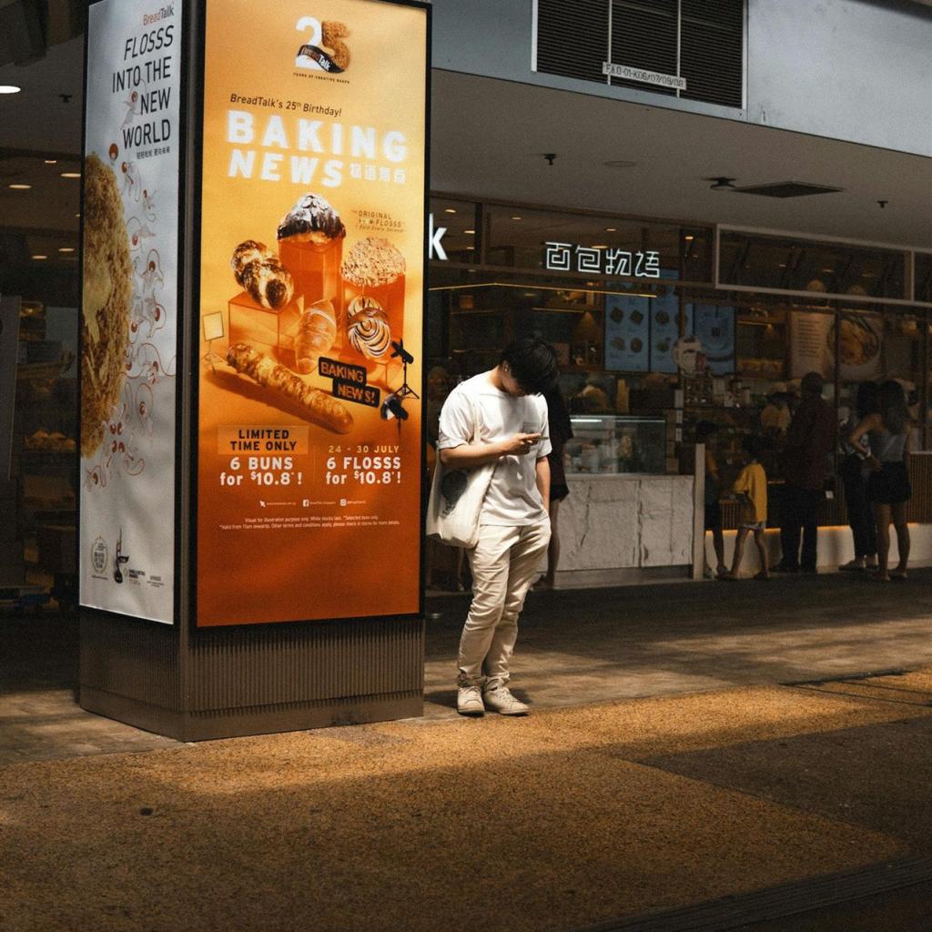 Ein Mann steht im Sonnenlicht neben einer Bäckereiwerbung in einer geschäftigen städtischen Umgebung.